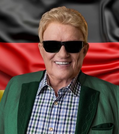 Der Sänger Heino mit blonder Frisur und schwarzer Sonnenbrille lächelt vor einer wehenden Deutschlandflagge. Er trägt eine grüne Trachtenjacke über einem blau-weiß karierten Hemd.