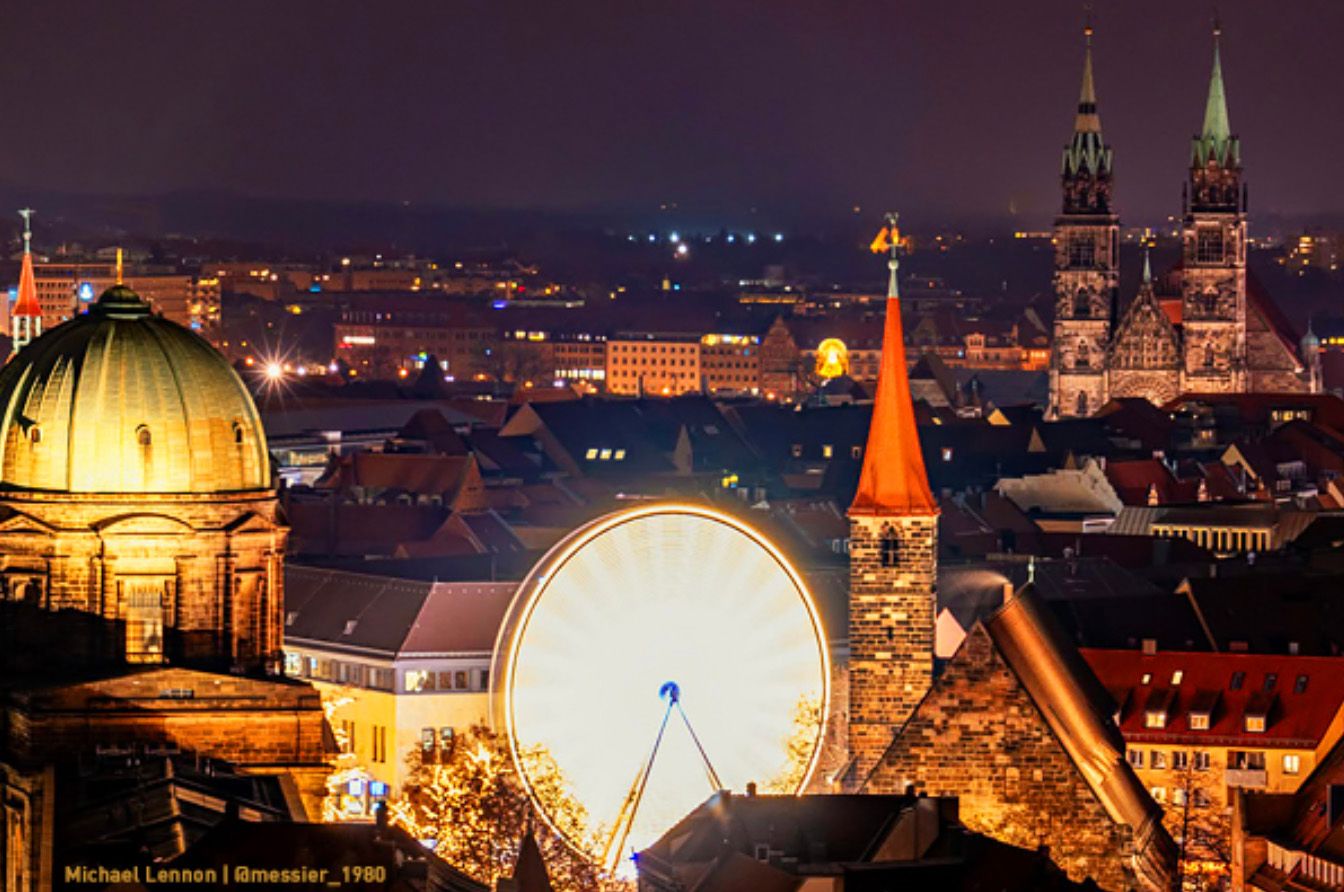 Blick bei Nacht über die Dächer der Nürnberger Altstadt auf das leuchtende Riesenrad vom WinterDorf Nürnberg, mit den Türmen der Lorenzkirche im Hintergrund.