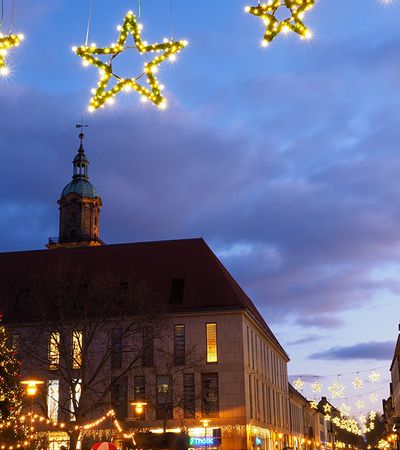 Weihnachtliche Beleuchtung in der Erlanger Innenstadt mit leuchtenden Sternen über den Straßen und einem geschmückten Tannenbaum vor historischen Gebäuden bei Abenddämmerung.