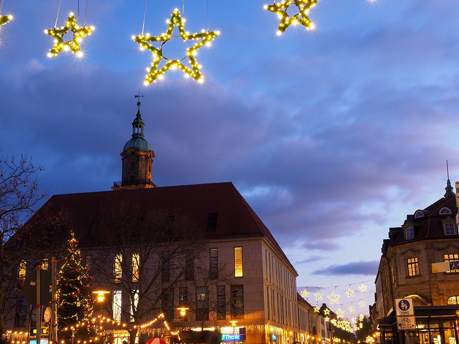 Weihnachtliche Beleuchtung in der Erlanger Innenstadt mit leuchtenden Sternen über den Straßen und einem geschmückten Tannenbaum vor historischen Gebäuden bei Abenddämmerung.