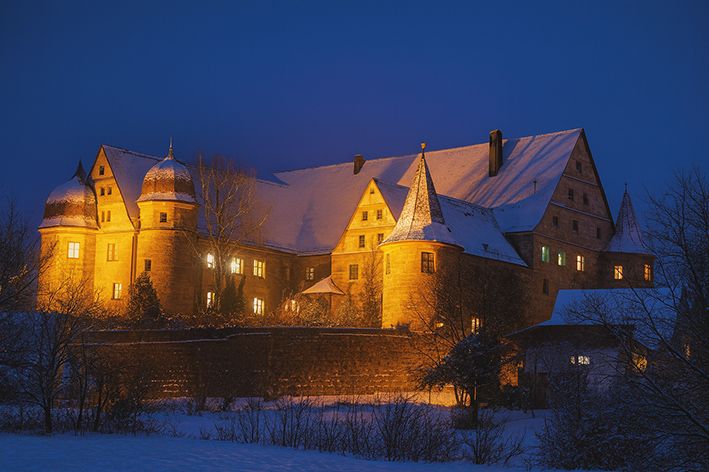 Das Schloss Wiesenthau an einem verschneiten Winterabend, warm angestrahlt, mit funkelnden Fenstern und tiefblauem Himmel – stimmungsvolle Kulisse für den Kunst- und Weihnachtsmarkt.