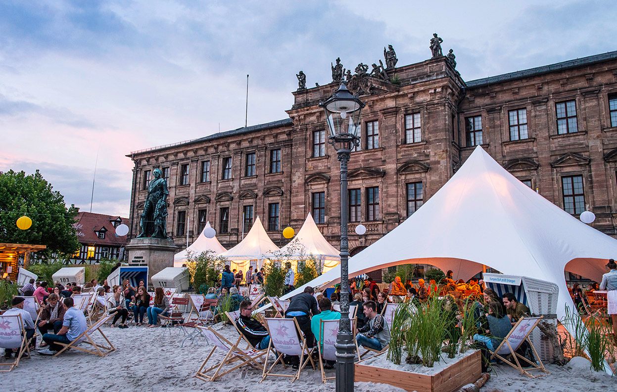 Besucher genießen den Sternla SchlossStrand 2026 vor dem Schloss Erlangen mit Sandstrand, Liegestühlen und sommerlicher Beachclub-Atmosphäre