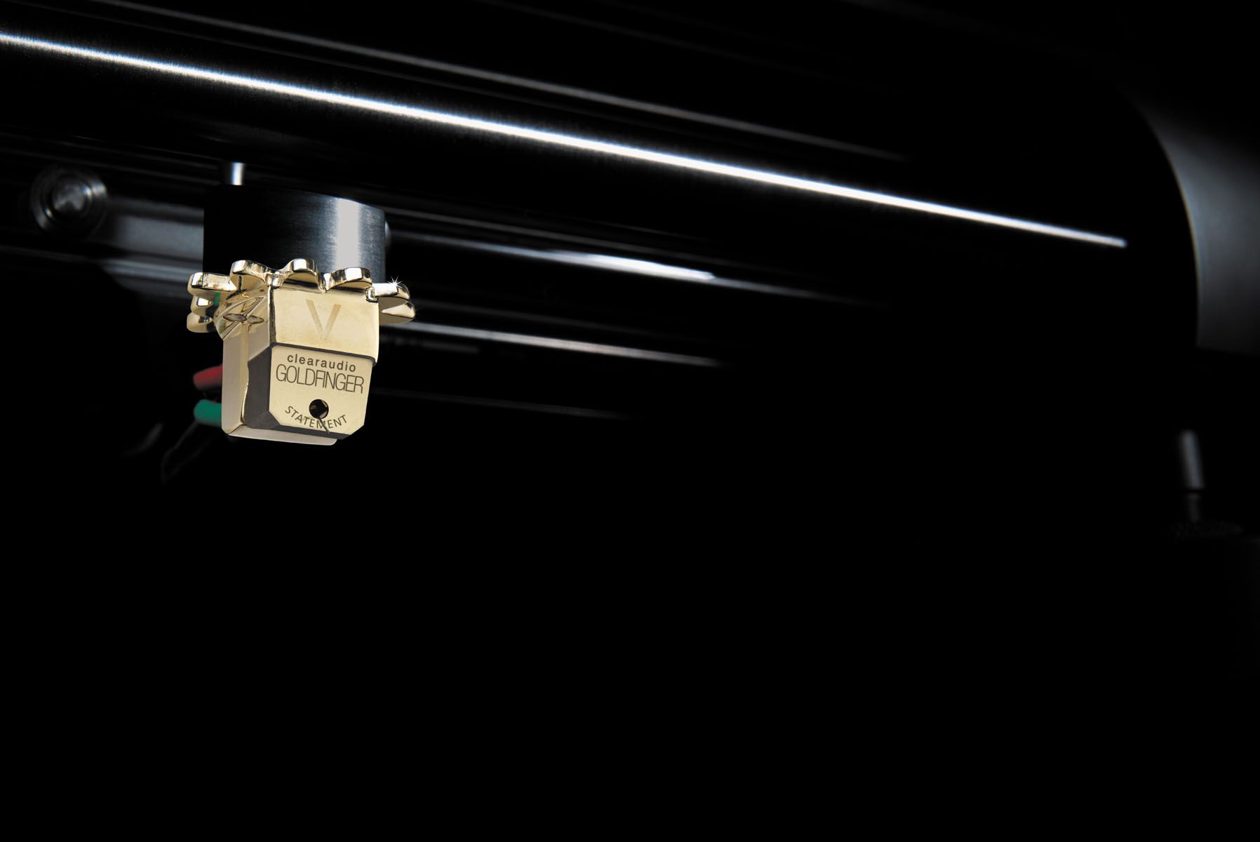 Macro shot of the golden clearaudio Goldfinger V2 cartridge mounted beneath a black tonearm in an atmospherically dimmed studio.