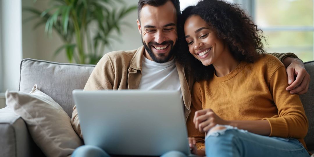 Smiling multiracial couple watches computer laptop sitting on sofa at home. Diverse husband, wife using pc online services. Technology lifestyle concept. Booking tickets shopping online.
