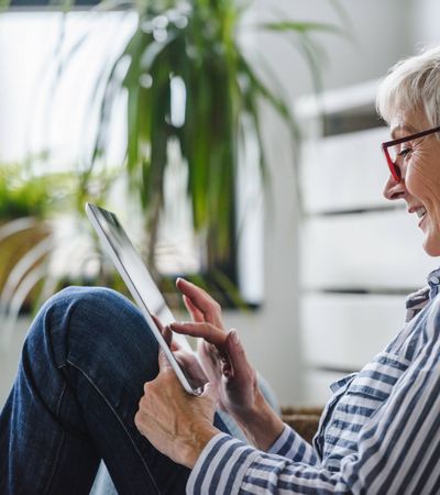 Eine kurzhaarige, ältere Frau mit Brille sitzt auf dem Fußboden und abonniert auf ihrem Tablet den mein PLATZ-Newsletter