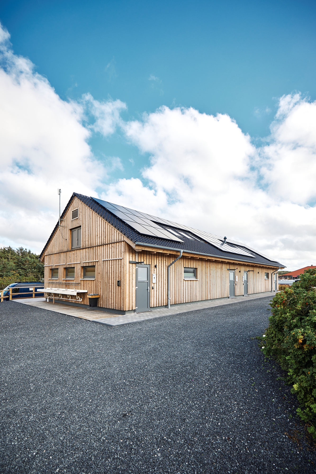 Modernes, langgezogenes Sanitärgebäude in Holzbauweise auf dem Campingplatz Westerland. Das Haus hat ein dunkles Satteldach mit großen Solarpanels, mehrere Türen und Fenster sowie Waschbecken an der Außenwand. Der Himmel ist leicht bewölkt, im Hintergrund sind Bäume und ein Wohnmobil sichtbar, im Vodergrund der Grandweg.