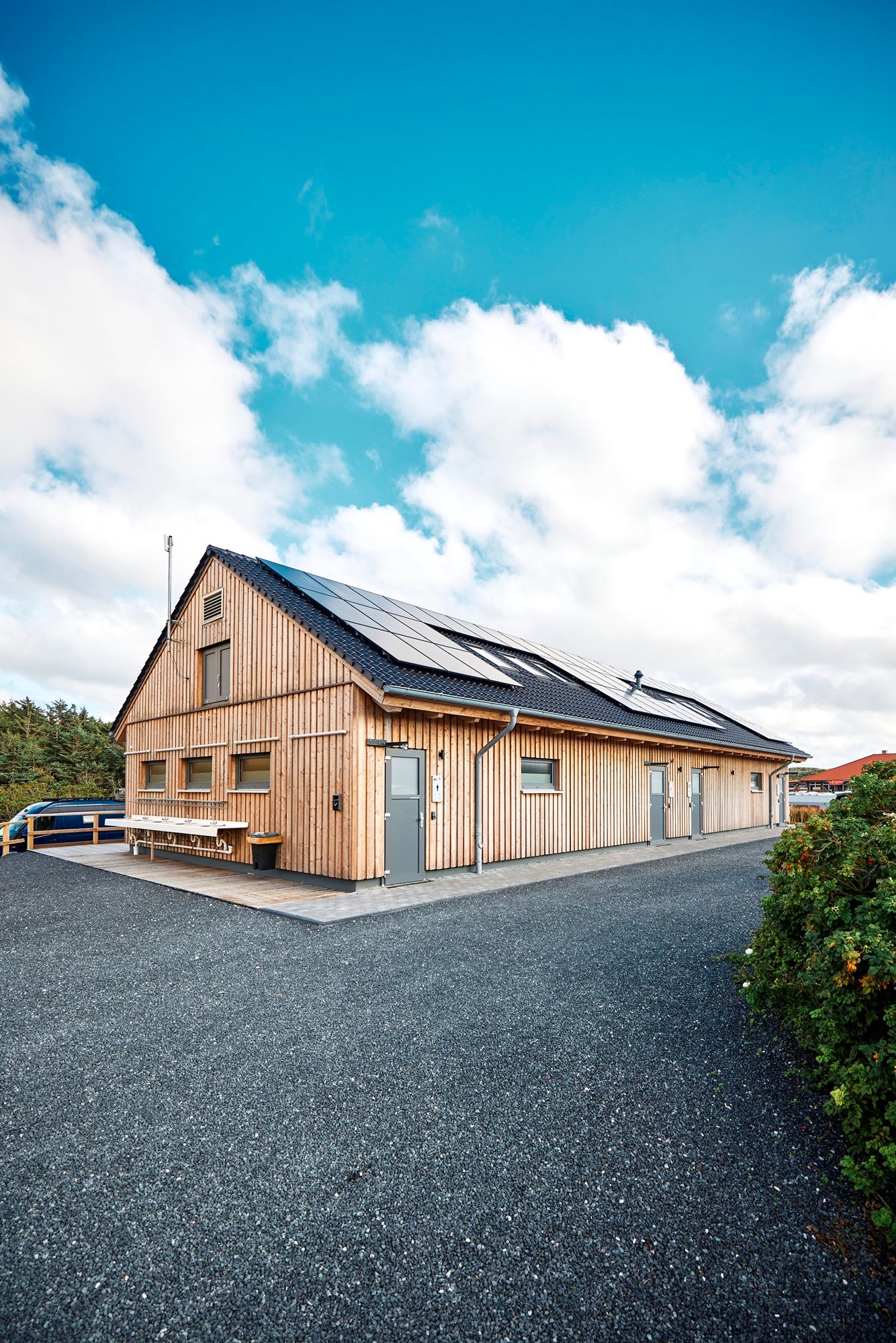 Modernes, langgezogenes Sanitärgebäude in Holzbauweise auf dem Campingplatz Westerland. Das Haus hat ein dunkles Satteldach mit großen Solarpanels, mehrere Türen und Fenster sowie Waschbecken an der Außenwand. Der Himmel ist leicht bewölkt, im Hintergrund sind Bäume und ein Wohnmobil sichtbar, im Vodergrund der Grandweg.