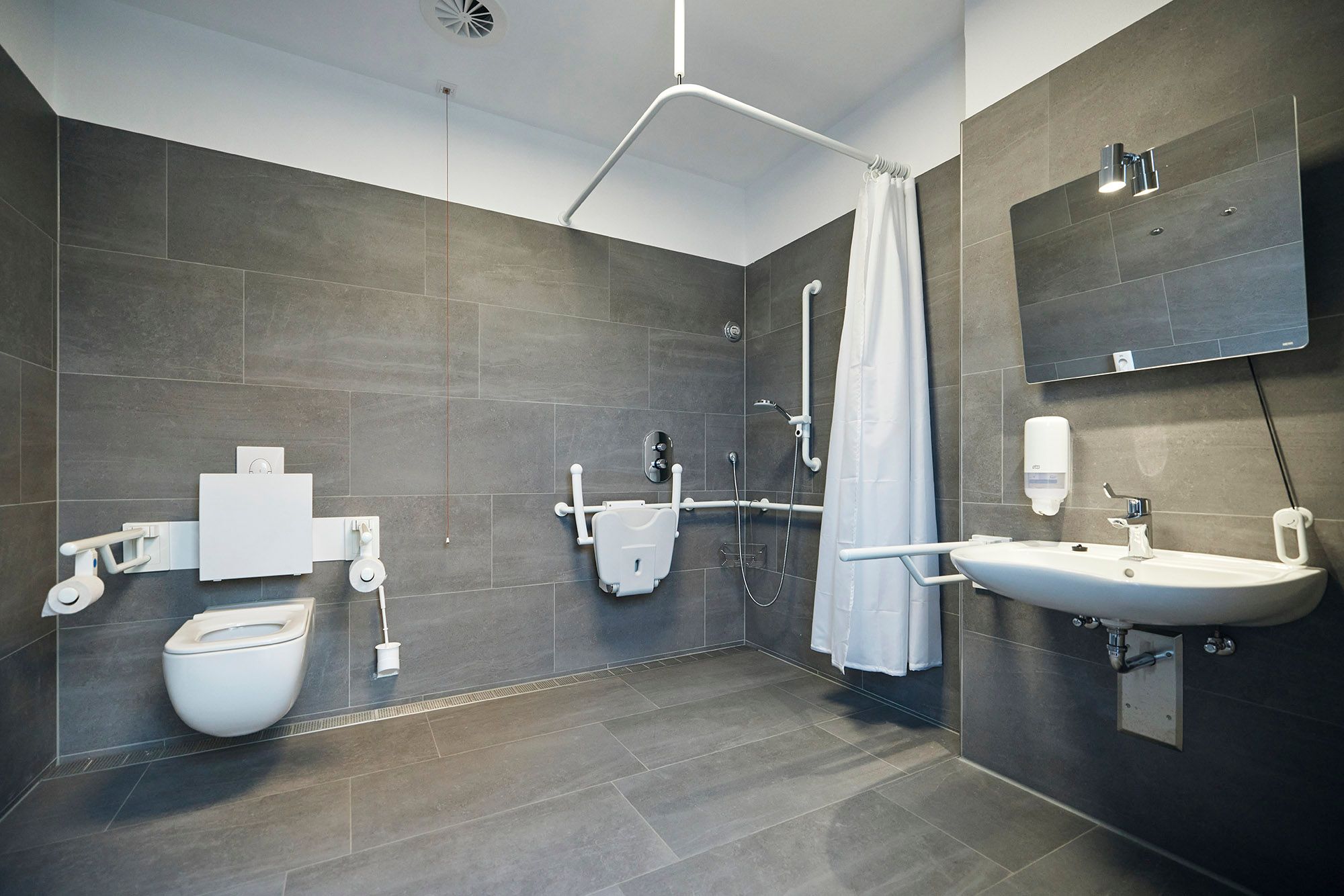 Rollstuhlgerechtes Badezimmer im Sanitärgebäude Süd auf dem Campingplatz Westerland - anthrazitfarbene Wand- und Bodenfliesen, weiße Badezeimmerkeramik.