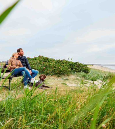 ein Paar sitzt auf einer Bank und blickt auf das Wattenmeer in Munkmarsch, zu ihren Füßen liegt ihr Hund