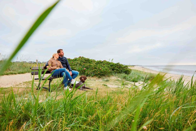ein Paar sitzt auf einer Bank und blickt auf das Wattenmeer in Munkmarsch, zu ihren Füßen liegt ihr Hund