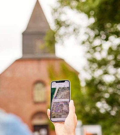 Eine Frau hält ihr Smartphone vor sich, um Informationen zur digitalen Gästeführung zur im Hintergrund sichtbaren Alten Dorfkirche zu erfassen.