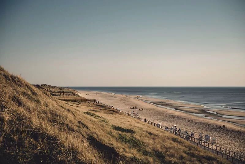 Blick von einem hohen Strandübergang in Westerland in Richtung Süden, man blickt auf die bewachsene Düne und sieht den Strand mit vereinzelten Strandkörben, die Nordsee sowie die Wandelbahn und zwei weitere Strandübergänge