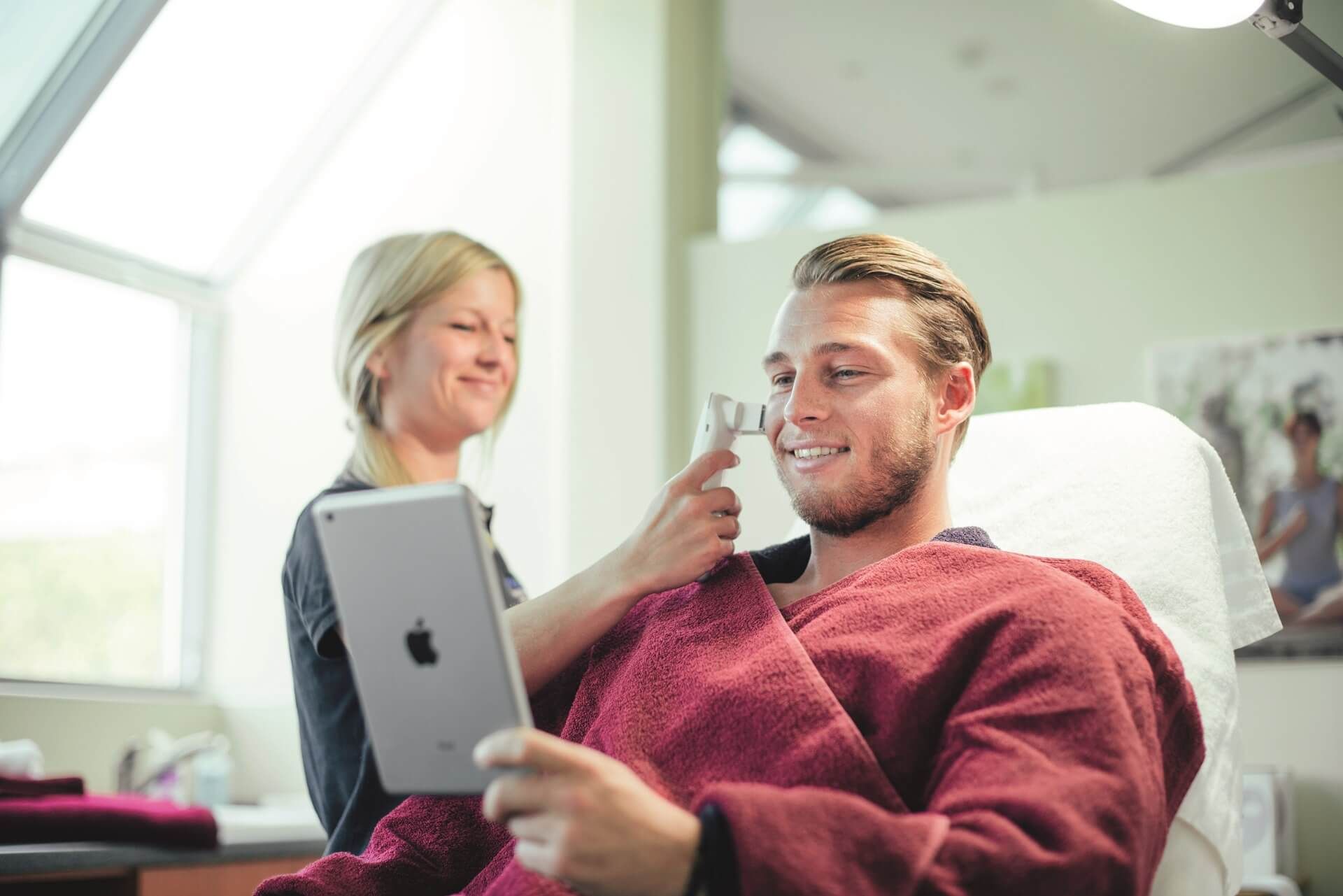 Mannträgt bordeauxroten Bademantel, erhält von einer Frau eine Hautanalyse und hält dabei ein iPad in der Hand.