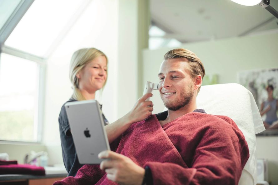 Mannträgt bordeauxroten Bademantel, erhält von einer Frau eine Hautanalyse und hält dabei ein iPad in der Hand.