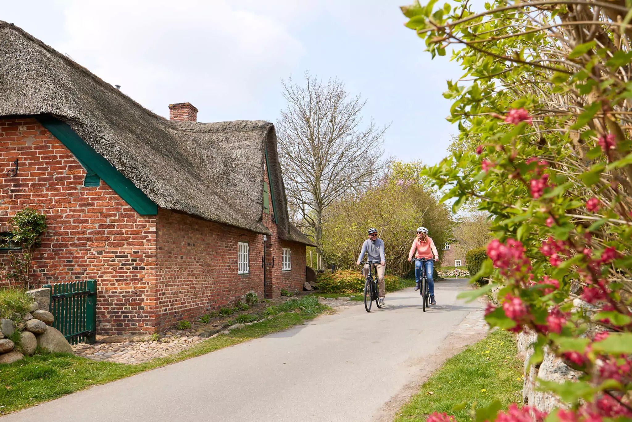 Frau und Mann fahren mit E-Bikes aus dem E-Mobility Center an einem Reetdachhaus in Keitum vorbei