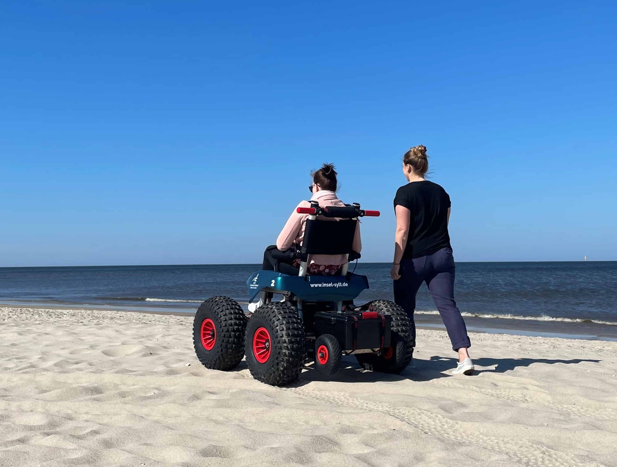 Elektrischer Strandrollstuhl am Westerländer Strand, nebenher spaziert eine weibliche Begleitung