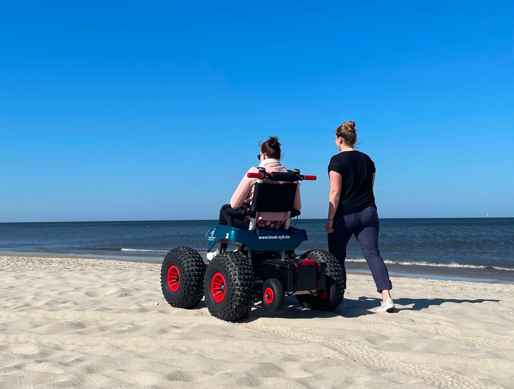 Elektrischer Strandrollstuhl am Westerländer Strand, nebenher spaziert eine weibliche Begleitung