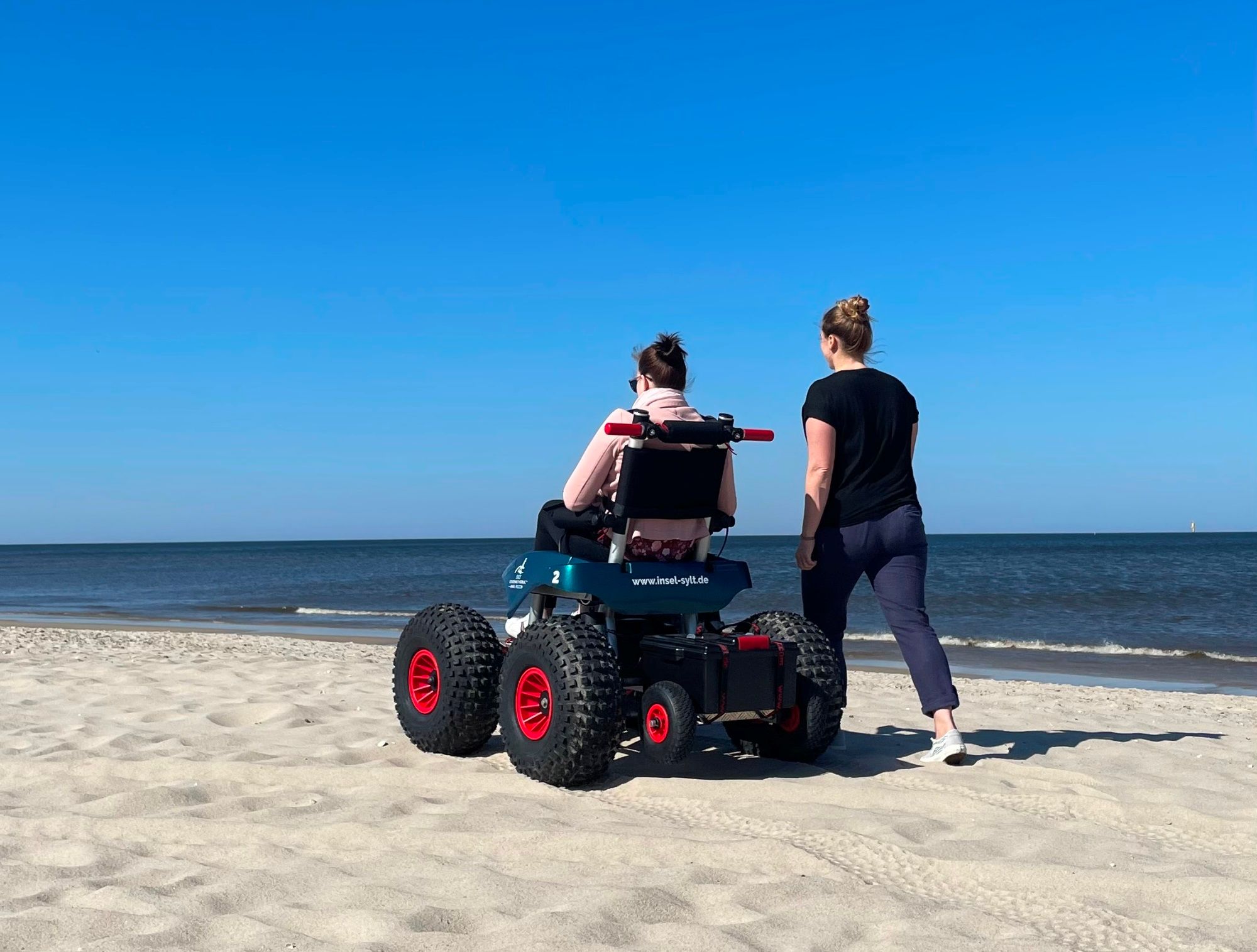 Elektrischer Strandrollstuhl am Westerländer Strand, nebenher spaziert eine weibliche Begleitung