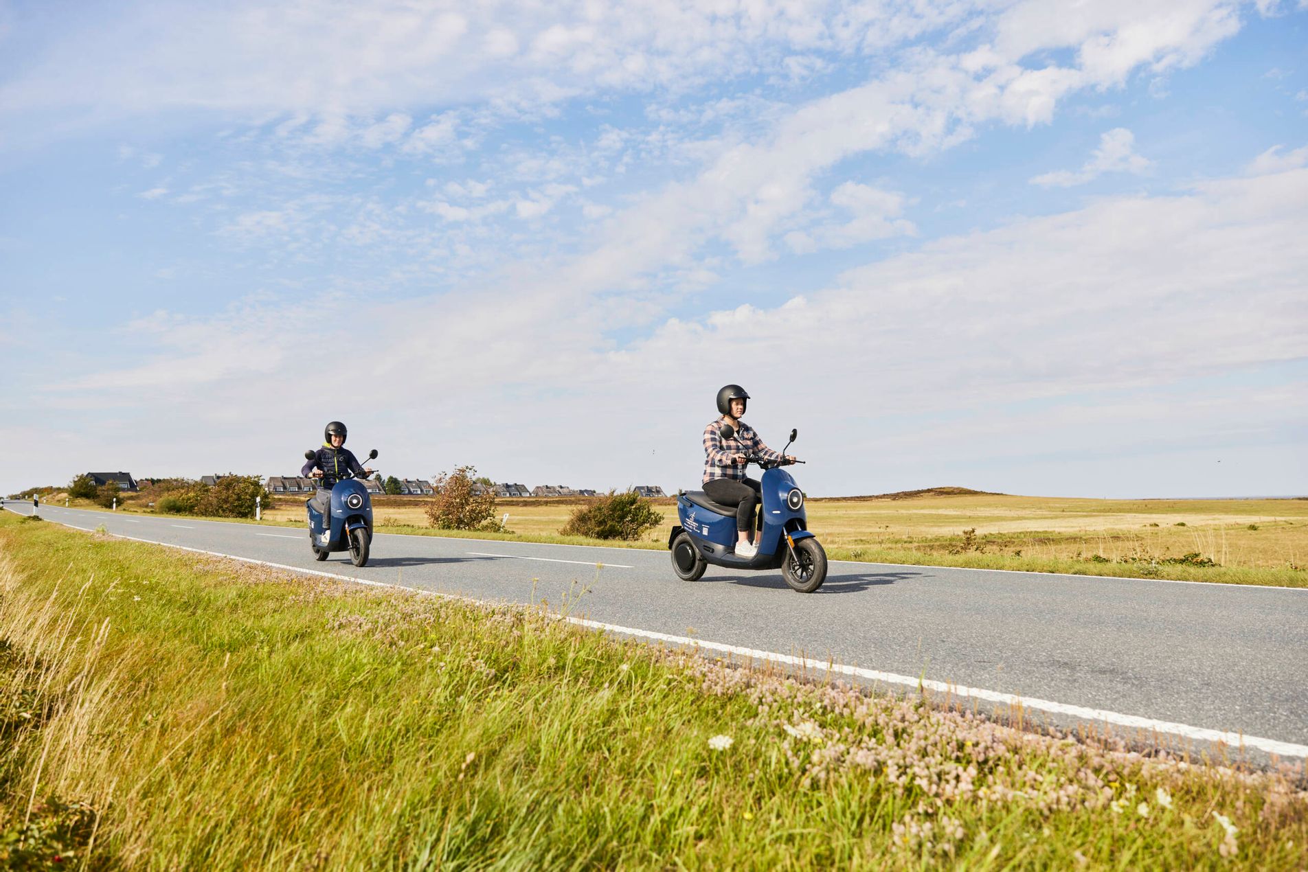 Zwei Frauen fahren mit blauen unu-Rollern über die Landstraße zwischen Keitum und Munkmarsch