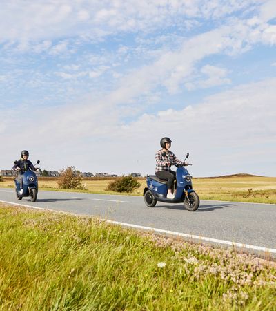 Zwei Frauen fahren mit blauen unu-Rollern über die Landstraße zwischen Keitum und Munkmarsch