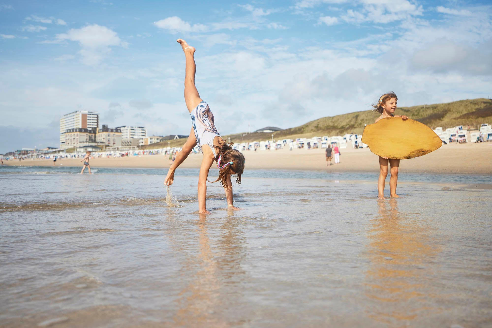 Mädchen macht Handstand im seichten Wasser der Nordsee während ein weiteres Mädchen mit Board im Wasser steht, im Hintergrund die Skyline von Westerland