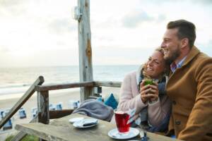 Frau kuschelt sich sitzend mit einem Becher Tee an einen neben ihr sitzenden Mann und blickt gemeinsam mit ihm auf die Nordsee