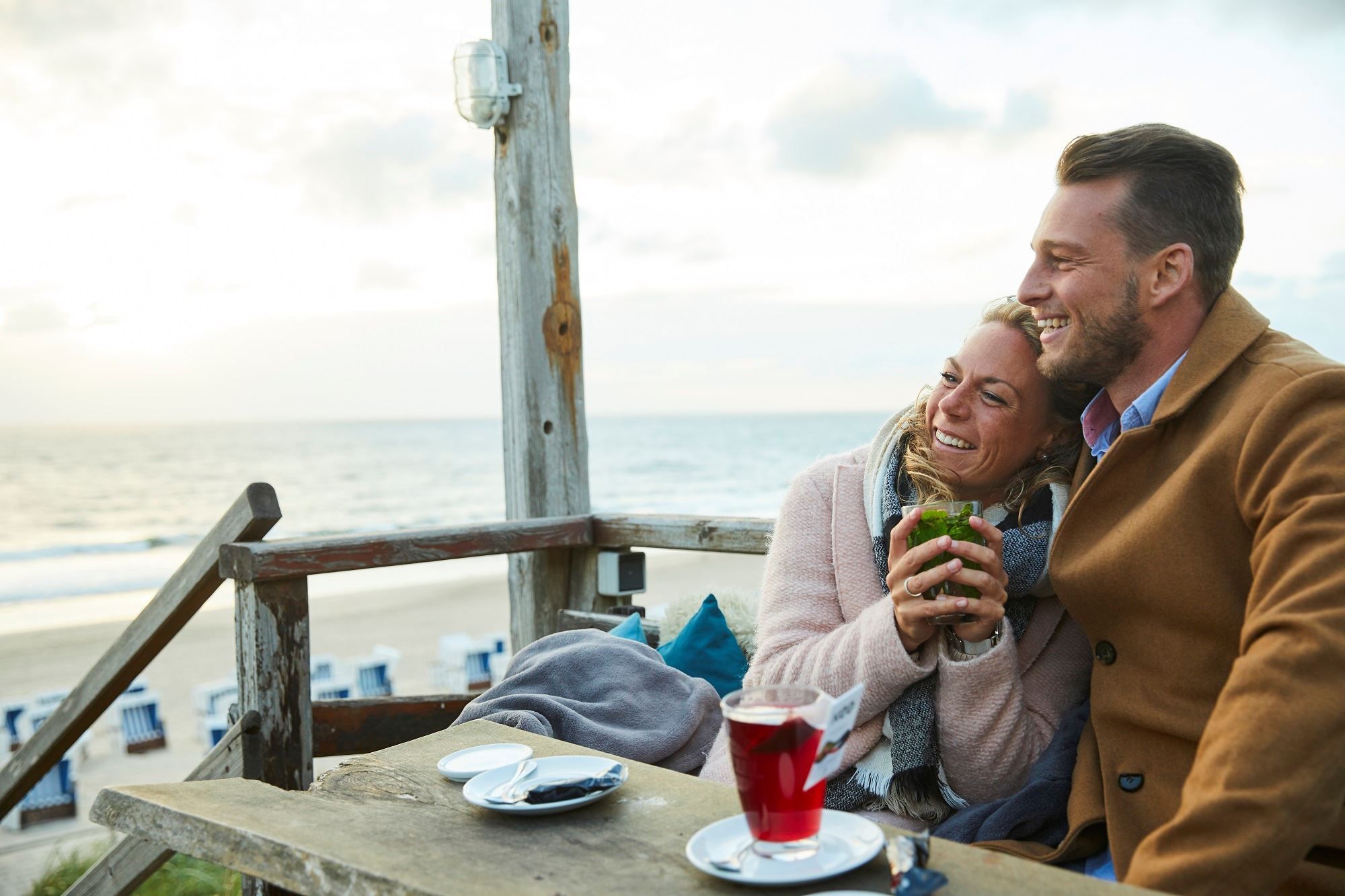 Frau kuschelt sich sitzend mit einem Becher Tee an einen neben ihr sitzenden Mann und blickt gemeinsam mit ihm auf die Nordsee