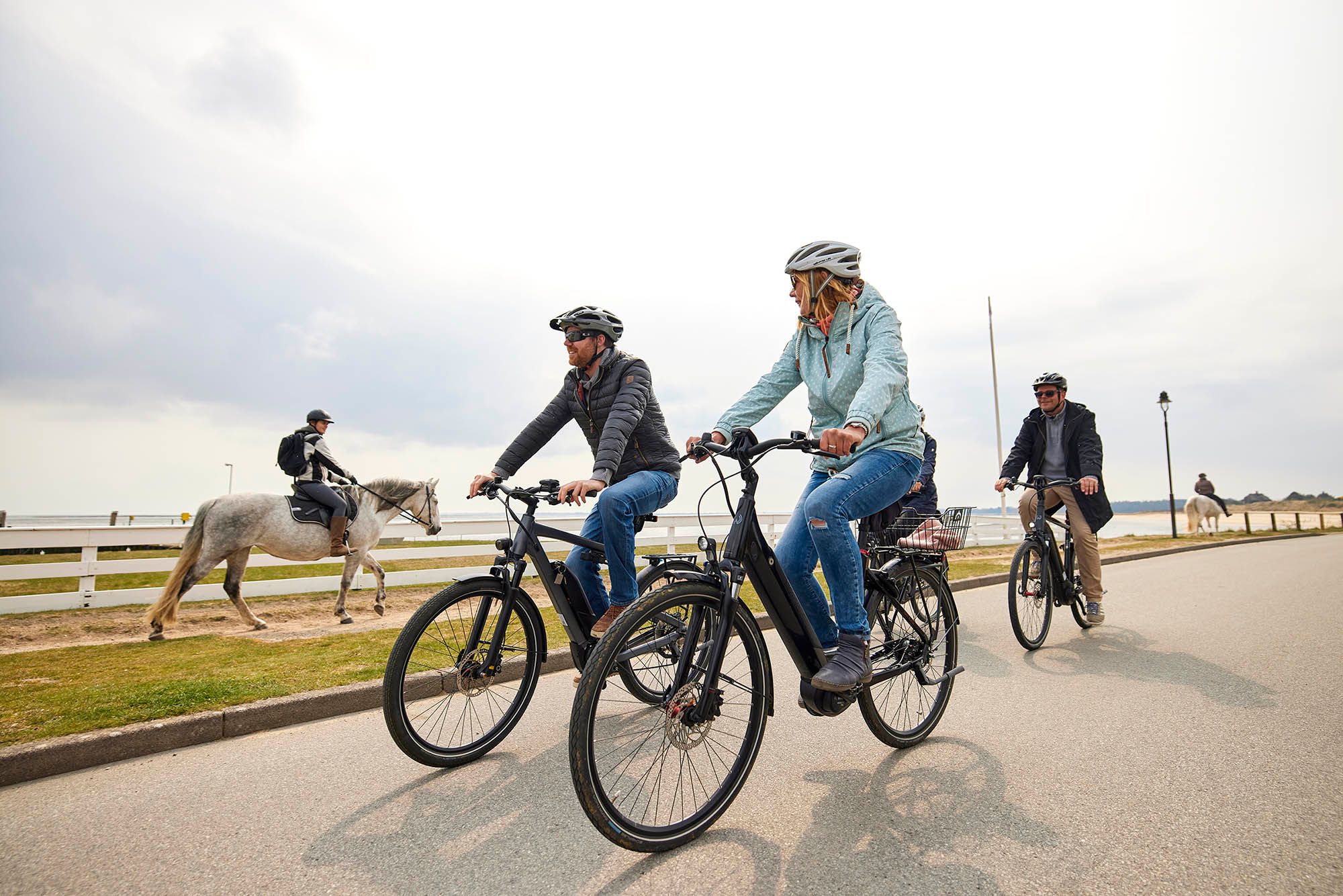 Zwei Männer und eine Frau fahren E-Bikes aus dem E-Mobility Center in Munkmarsch. Auf dem Reitweg neben ihnen werden zwei Schimmel geritten.