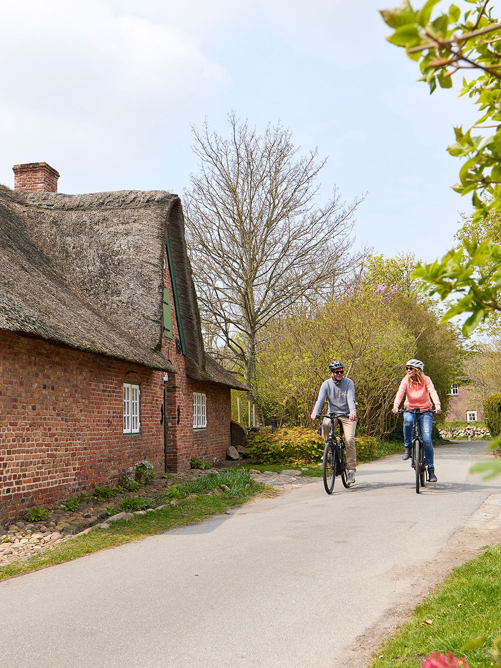 Frau und Mann fahren mit E-Bikes aus dem E-Mobility Center an einem Reetdachhaus in Keitum vorbei