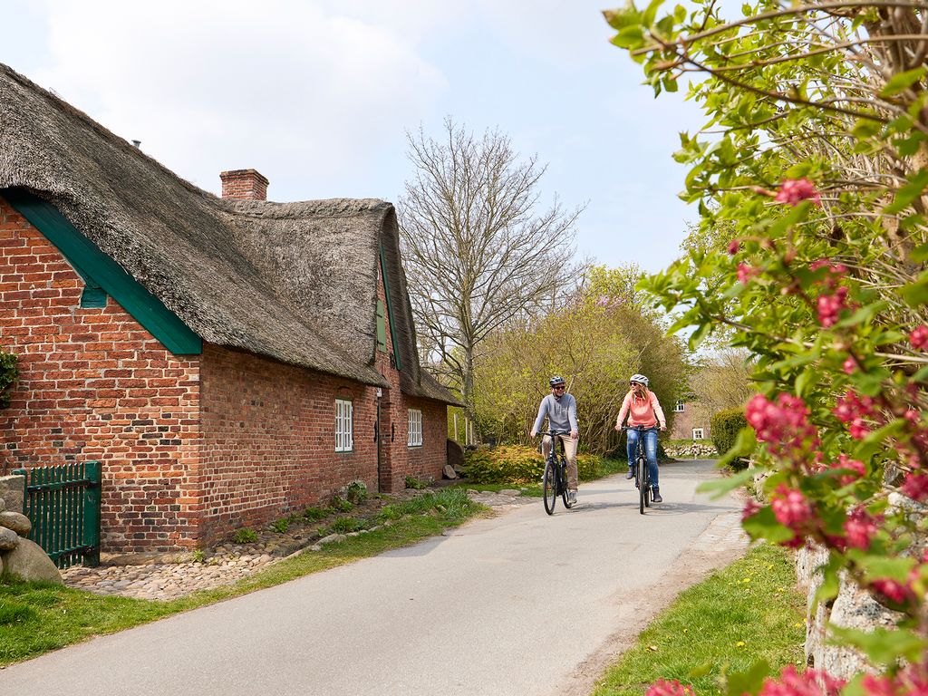 Frau und Mann fahren mit E-Bikes aus dem E-Mobility Center an einem Reetdachhaus in Keitum vorbei