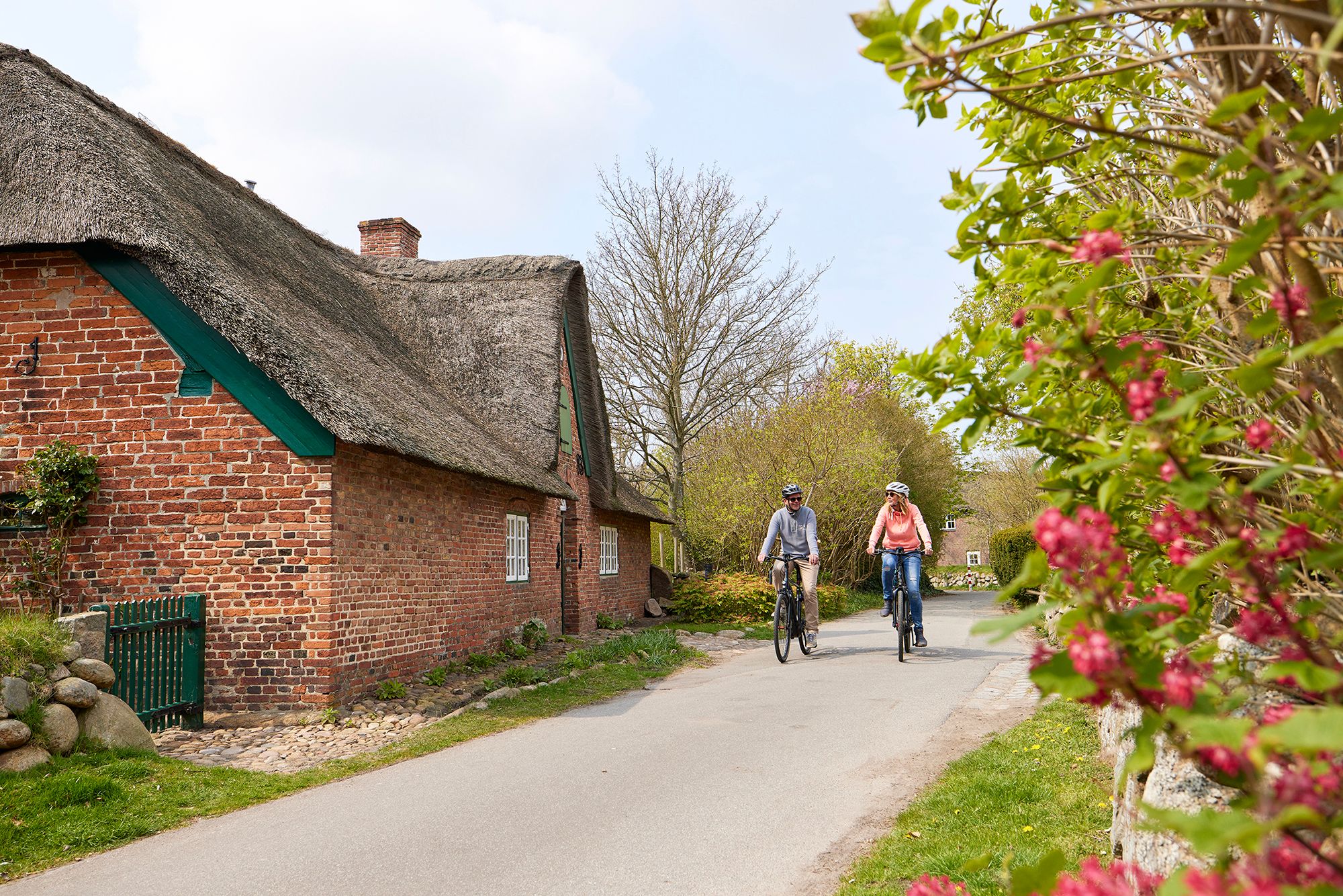 Frau und Mann fahren mit E-Bikes aus dem E-Mobility Center an einem Reetdachhaus in Keitum vorbei