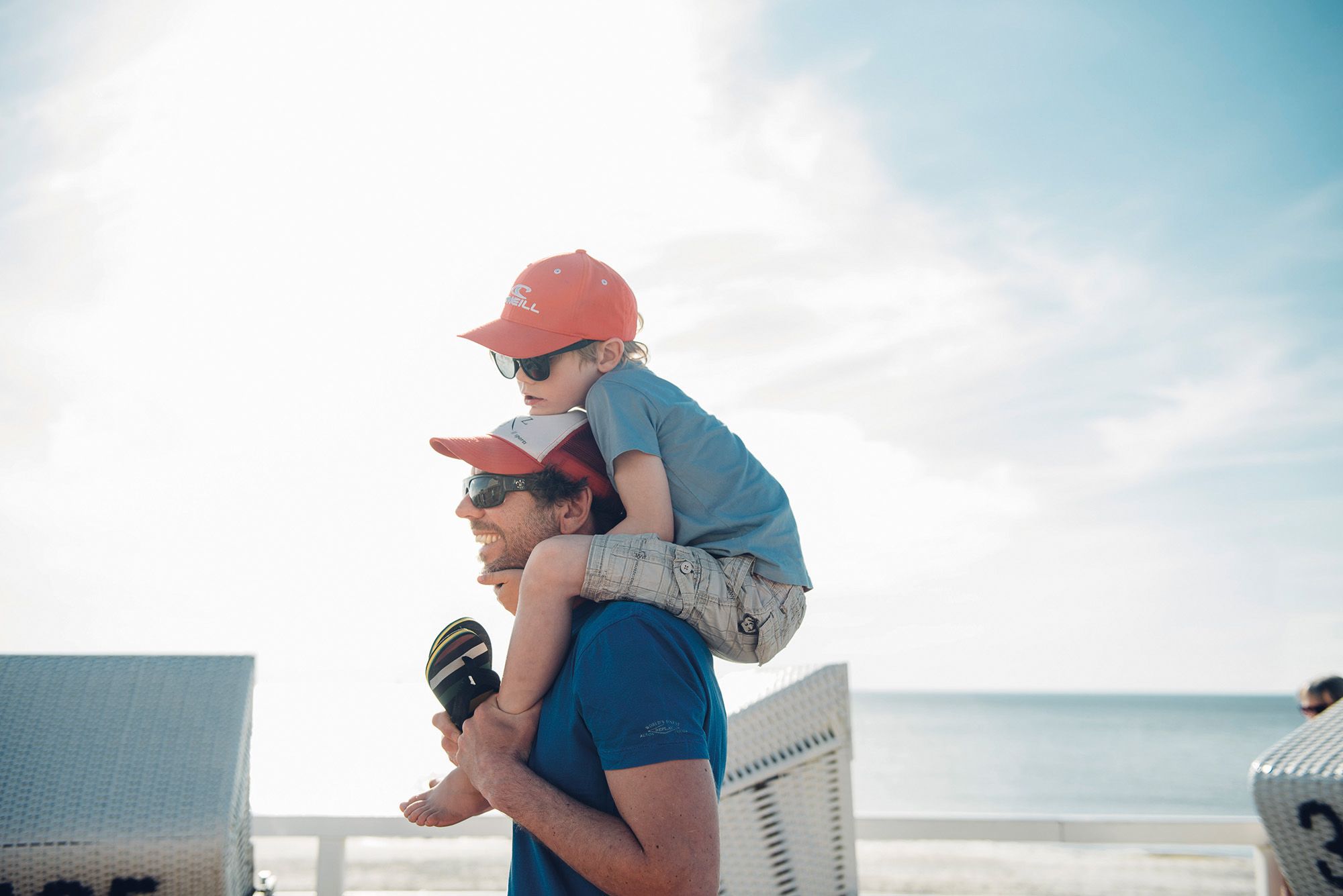 Mann trägt einen Jungen auf den Schultern über die Promenade in Westerland auf Sylt, im Hintergund die Nordsee bei Sonnenschein