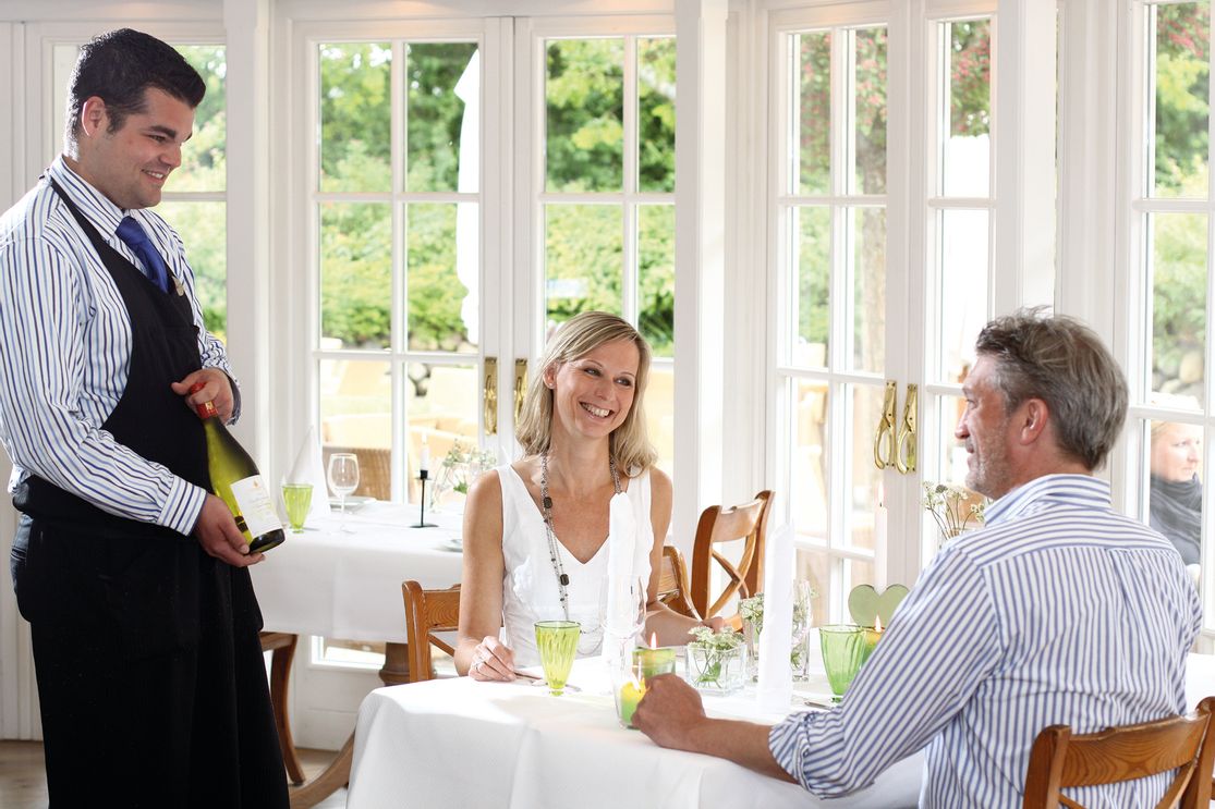Mann und Frau sitzen an einem Tisch in einem Restaurant in Keitum und bekommen von einem Kellner eine Flasche Wein gezeigt
