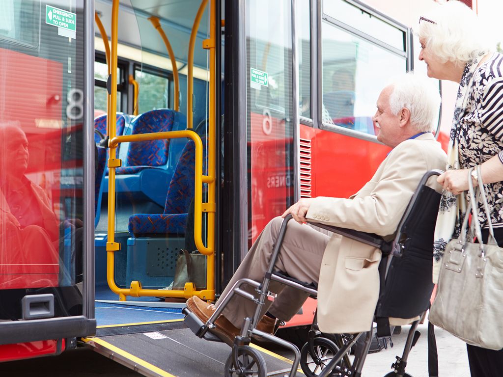 Seniorin schiebt Senior im Rollstuhl in einem rotfarbenen Linienbus