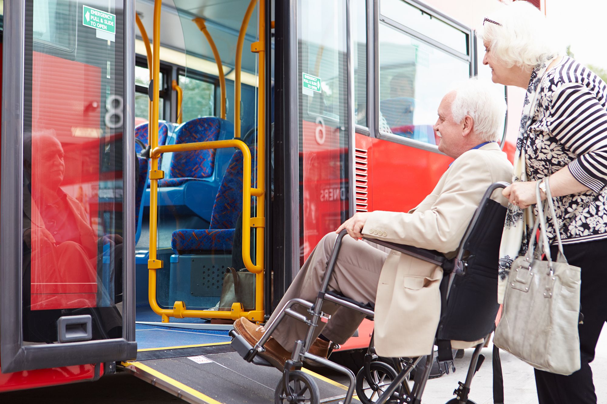 Seniorin schiebt Senior im Rollstuhl in einem rotfarbenen Linienbus