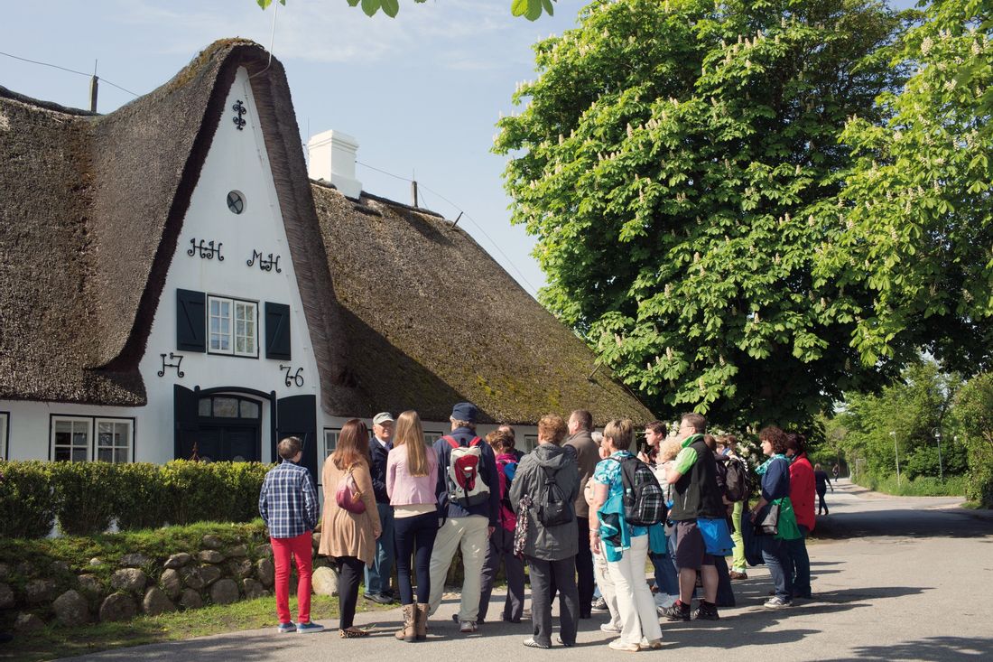 Teilnehmer an der Ortsführung durch Keitum stehen vor einem weißen Reetdachhaus und lauschen dem Gästeführer