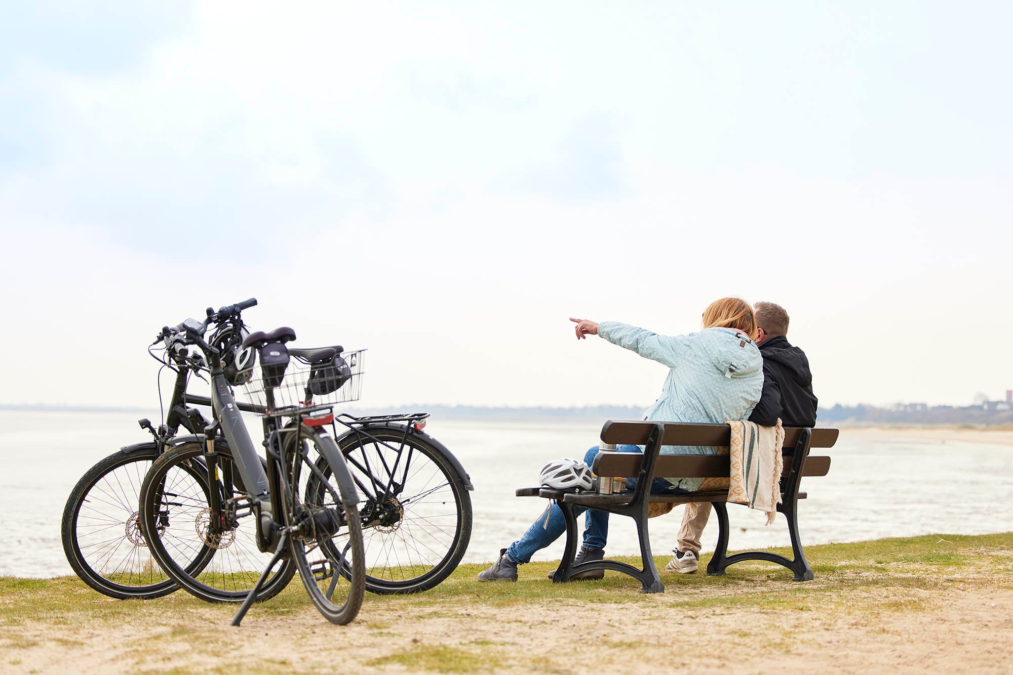 Zwei Personen sitzen in Munkmarsch auf einer Bank, neben Ihnen zwei E-Bikes aus dem E-Mobility Center und blicken auf´s Wattenmeer