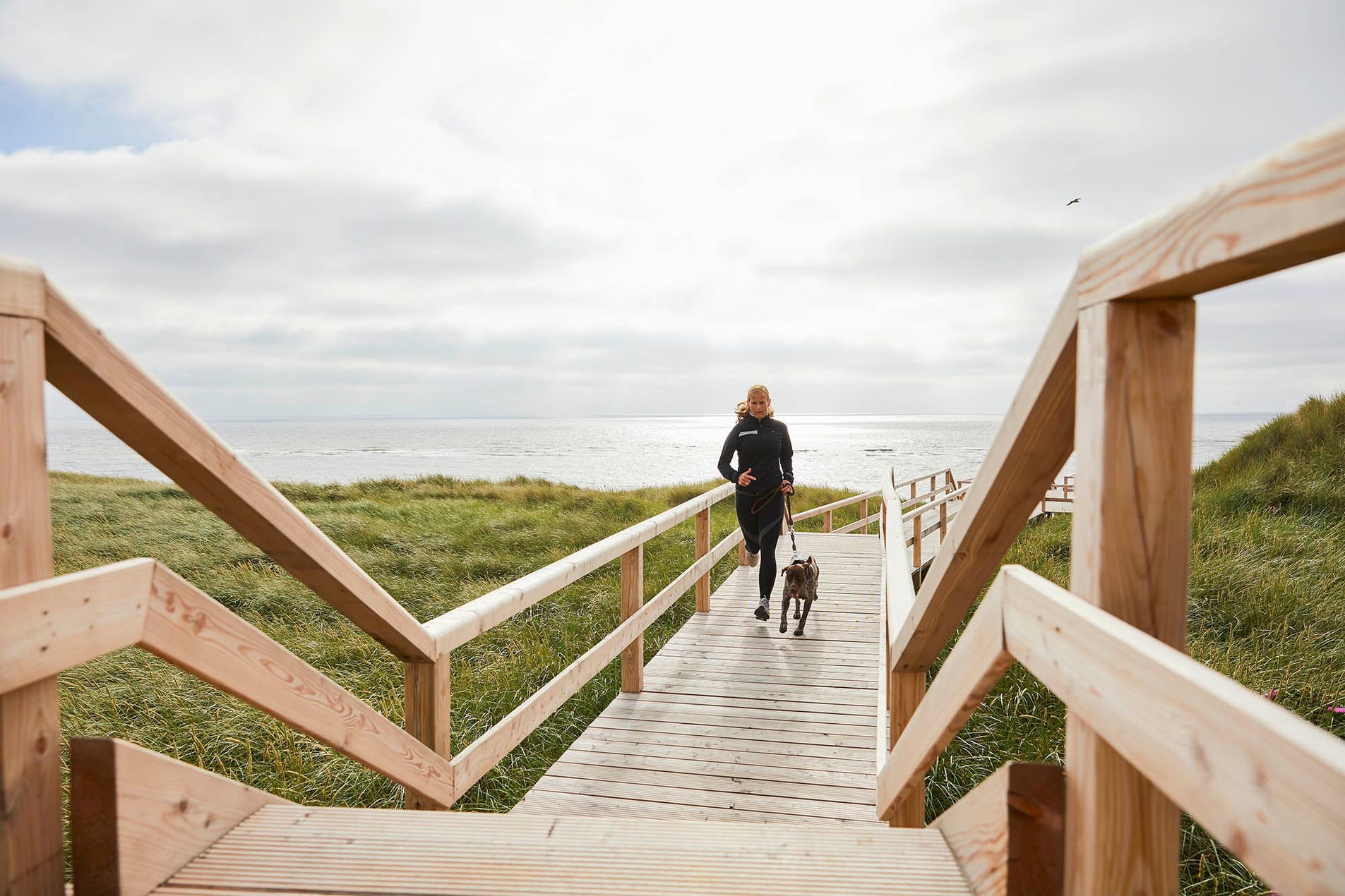 Joggerin mit Hund auf einem Strandübergangssteg auf Sylt