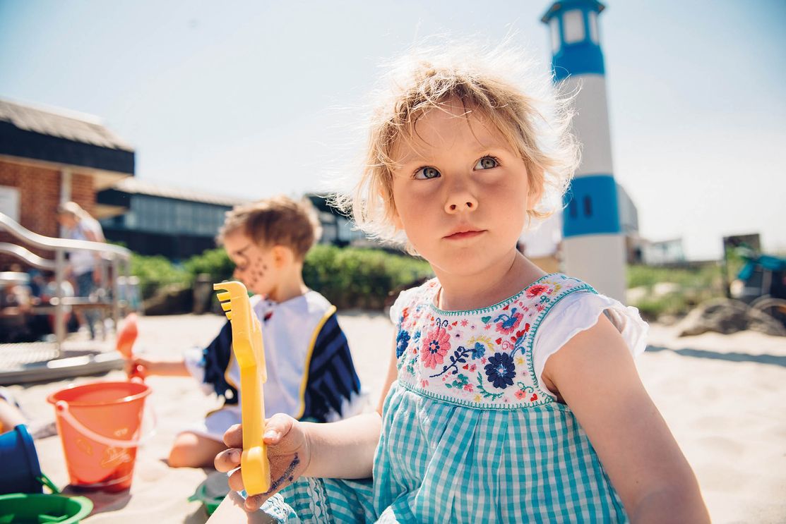 Kleines Mädchen hat auf dem Spielplatz an der Villa Kunterbunt in Westerland Sandspielzeug in der Hand und schaut an der Kamera vorbei. Im Hintergrund sitzt ein kleiner Junge mit im Sand