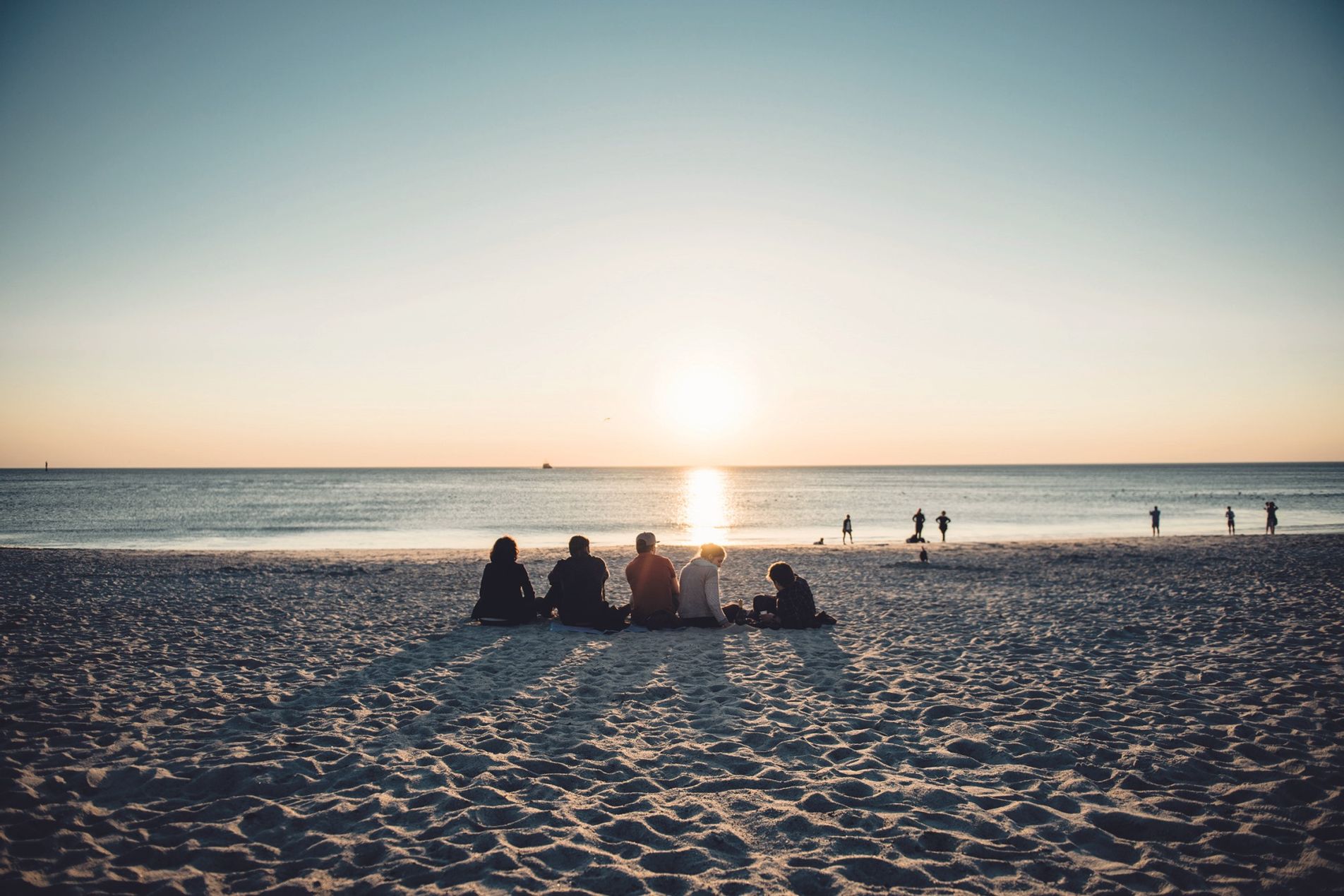 Gruppe von mehreren Menschen sitzt mit dem Rücken zur Kamera am Nordseestrand von Sylt und blickt auf den Sonnenuntergang