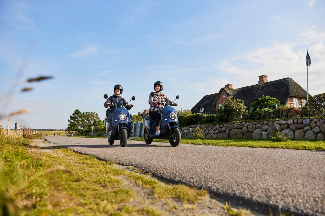 Zwei weibliche Personen fahren mit einen unu E-Roller aus dem E-Mobility Center vor einem Friesenhaus in Keitum auf Sylt