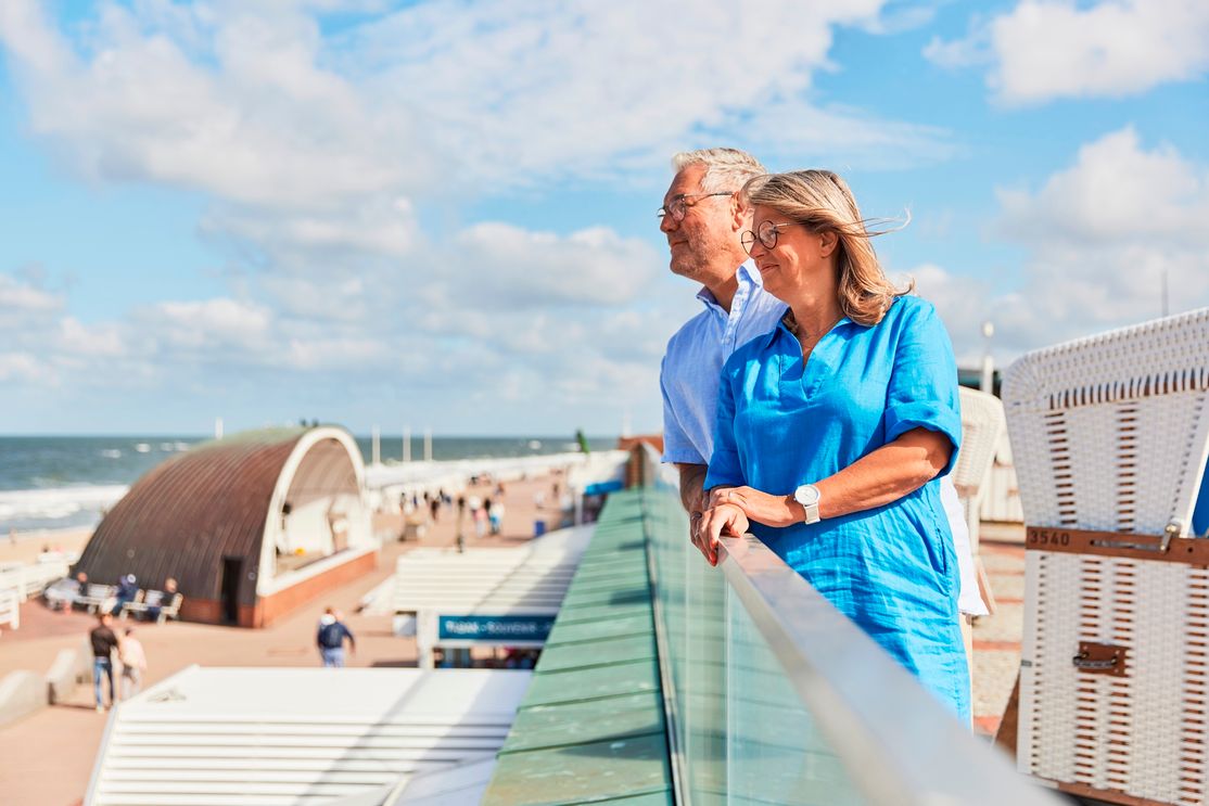 Paar steht auf der oberen Strandpromenade in Westerland und blickt auf die Nordsee, im Hintergrund ein Strandkorb und die Musikmuschel