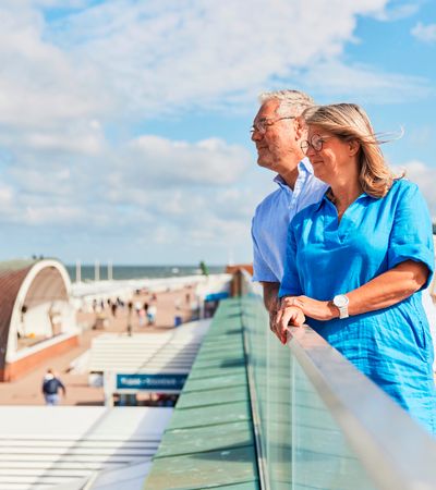 Paar steht auf der oberen Strandpromenade in Westerland und blickt auf die Nordsee, im Hintergrund ein Strandkorb und die Musikmuschel