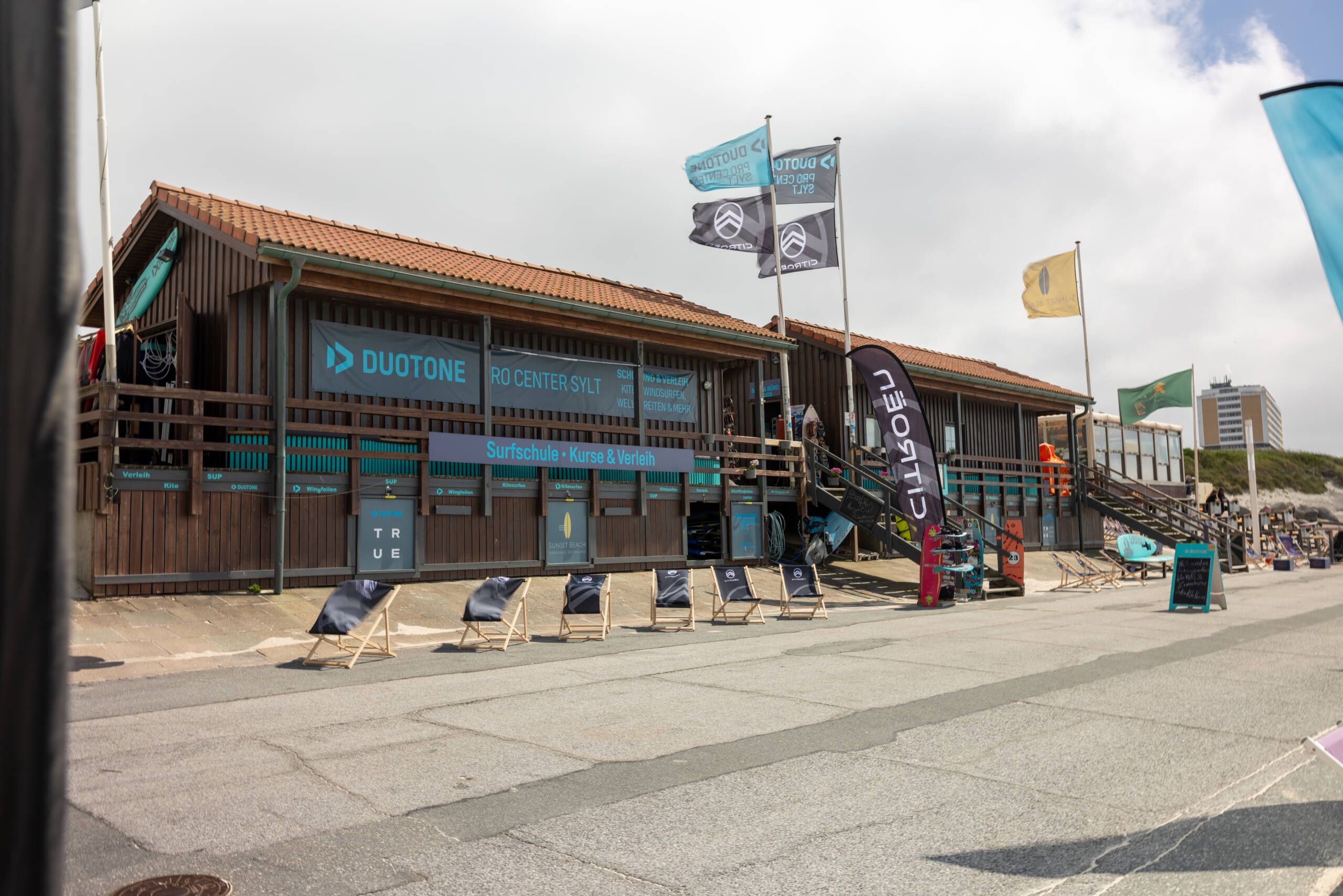 Surfschule mit Holzgebäude und mehreren Fahnen, vor dem Liegestühle aufgereiht sind.