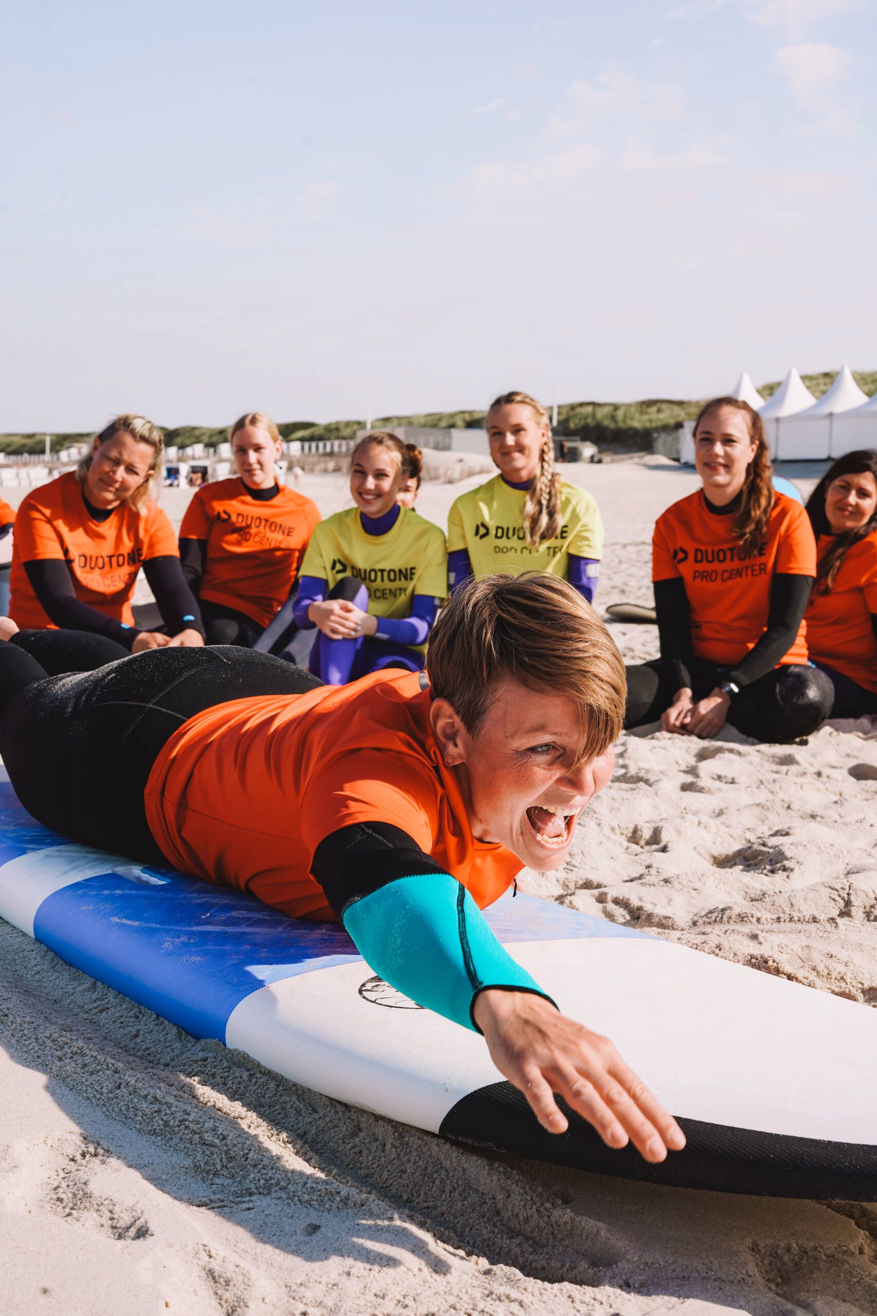 Frau, die auf einem Surfbrett im Sand liegt mit Schülern, die zuschauen.
