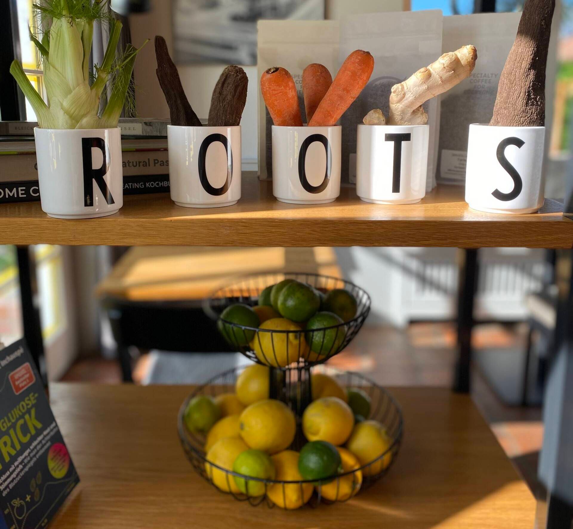 Ein Regal mit einer Schale Obst und darüber fünf Tassen aufgestellt mit je einem Buchstaben drauf, welche das Wort Roots bilden.