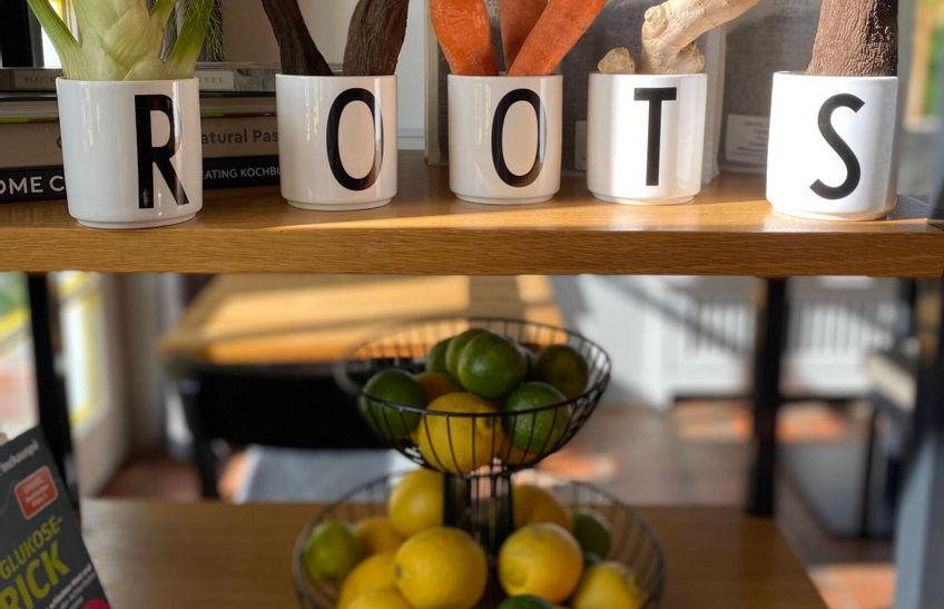 Ein Regal mit einer Schale Obst und darüber fünf Tassen aufgestellt mit je einem Buchstaben drauf, welche das Wort Roots bilden.