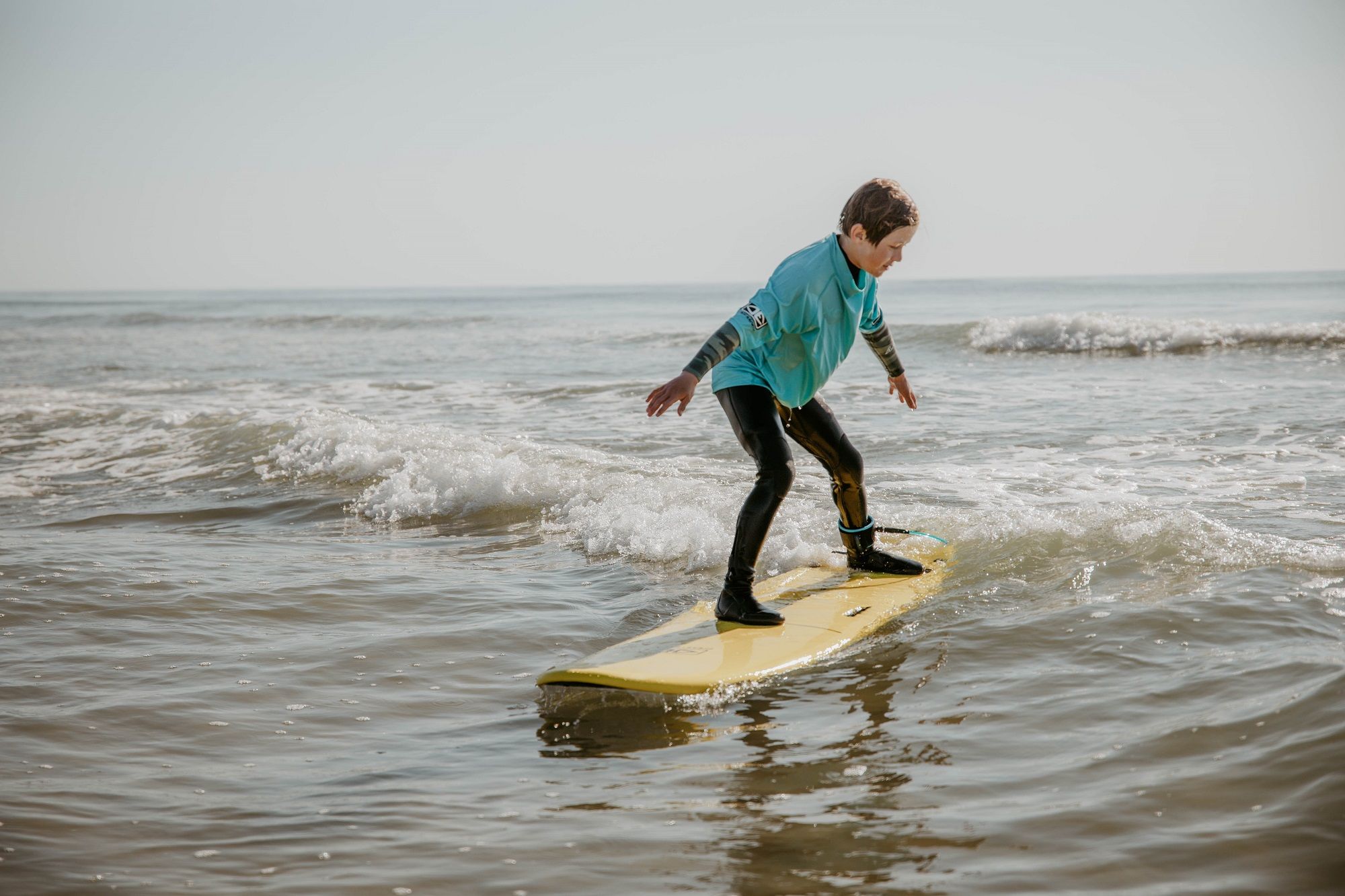 Ein Kind steht auf einem gelben Surfbrett und reitet eine kleine Welle im flachen Wasser. Es trägt ein türkisfarbenes Surfshirt über einem Neoprenanzug sowie schwarze Surfschuhe. Die Arme sind zur Balance ausgestreckt, der Blick ist konzentriert nach vorn gerichtet. Im Hintergrund ist das ruhige Meer unter klarem Himmel zu sehen.