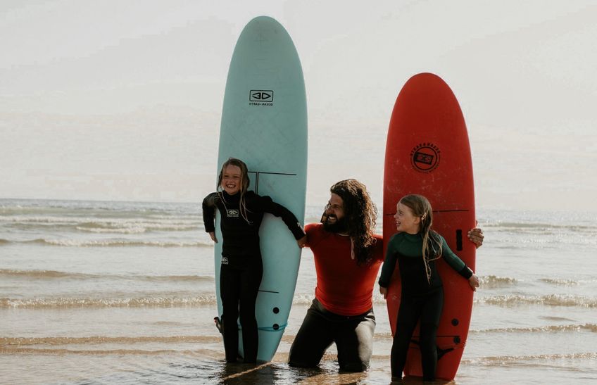 Ein Mann kniet im seichten Wasser am Strand und lacht gemeinsam mit zwei Kindern, die jeweils ein Surfbrett halten. Das Kind links steht hinter einem blauen Surfbrett, das Kind rechts lehnt sich an ein rotes Surfbrett. Alle tragen Neoprenanzüge und im Hintergrund ist das ruhige Meer zu sehen.