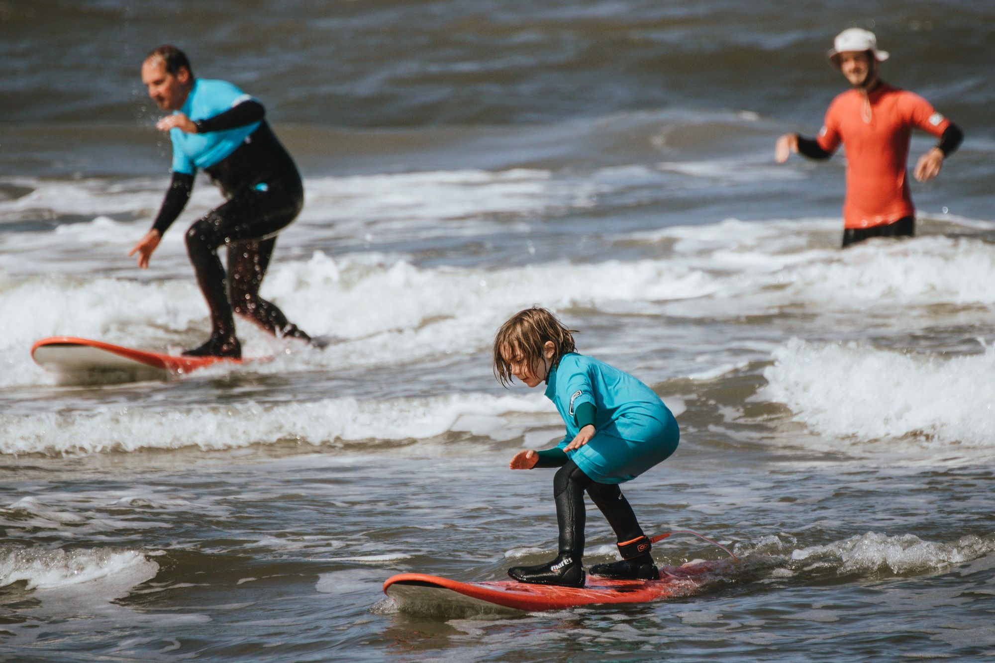 Ein Kind im Vordergrund surft auf einem kleinen Wellengang und balanciert auf einem roten Surfbrett. Es trägt einen türkisfarbenen Neoprenanzug und schwarze Surfschuhe. Im Hintergrund sind ein erwachsener Mann auf einem Surfbrett und ein weiterer Mann im Wasser zu sehen, beide in Neoprenanzügen. Die Szene spielt im flachen Wasser am Meer bei leichter Brandung.